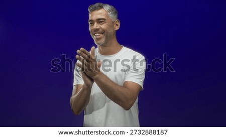 Man middleaged clapping hands in studio wearing white tshirt, smiling broadly toward camera; happiness celebration applause.
