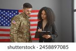 A man in military uniform and a businesswoman with a clipboard confer in front of an american flag in an office.