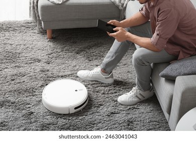 Man with mobile phone and modern robot vacuum cleaner sitting on sofa at home