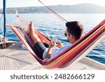 Man with mobile phone relaxing on yacht in hammock. Sea travel on sailboat. Traveler gaming on smartphone in summer vacation. Concept of doomscrolling, digital wellbeing, surfing the Internet, FOMO