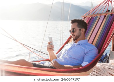 Man with mobile phone relaxing on yacht in hammock. Sea travel on sailboat. Traveler using smartphone and mobile app for banking, shopping online in summer vacation. Digital wellbeing, lifestyle - Powered by Shutterstock