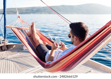 Man with mobile phone relaxing on yacht in hammock. Sea travel on sailboat. Traveler gaming on smartphone in summer vacation. Concept of doomscrolling, digital wellbeing, surfing the Internet, FOMO - Powered by Shutterstock