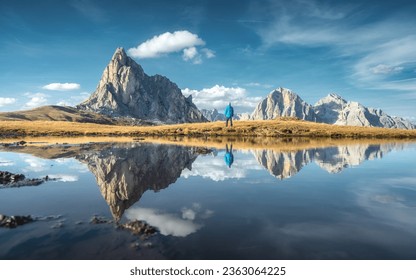 Man and mountains reflected in lake at sunset in autumn. Passo Giau, Dolomites, Italy. Standing guy on the shore of lake is looking on high rocks and blue sky with clouds in fall. Reflection in water - Powered by Shutterstock