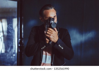A Man Moviemaker Shoots With a Vintage Camera. A Beard and a Black Jacket on it Makes it Solid. The Man is in a Dark Room.