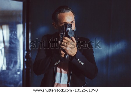 A Man Moviemaker Shoots With a Vintage Camera. A Beard and a Black Jacket on it Makes it Solid. The Man is in a Dark Room.