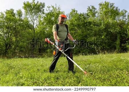 Man mowing the grass at his garden by using string trimmer with protective helmet and gloves.