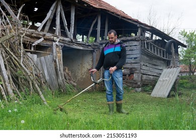 Man Mowing Tall Grass With Petrol Lawn Trimmer In The Village Rustic Yard. Gardening Care Equipment. Process Of Lawn Trimming With Hand Mower