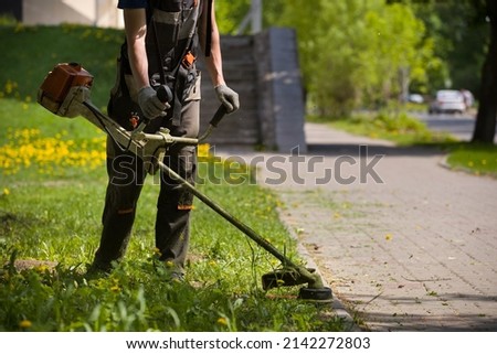 A man mows the grass in a grassy field along a footpath on a warm sunny day. A man in protective clothing, and gloves with a trimmer. 商業照片 © 