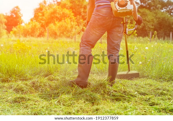 A man mows the grass with a hand-held lawn mower in\
the backyard in the sunlight. The gardener mows the long grass on\
the lawn in the garden with a trimmer. A farmer\'s lifestyle.\
Selective, soft focus