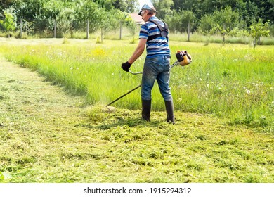 A man mows the grass with a trimmer on a sunny day. Back shot of a farmer mowing grass in the backyard. A quiet scene. A view from the back of a man with a gasoline mower.
