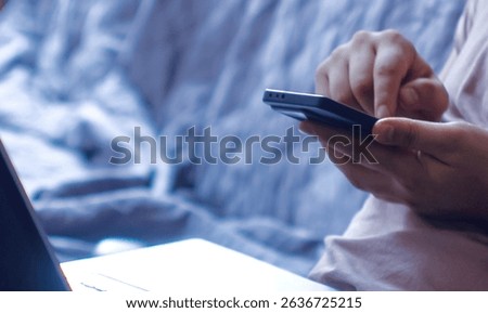 Man multitasking at home using smartphone and laptop in cozy interior. Close-up of hands holding smartphone and browsing, while laptop is open in the background.