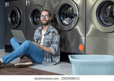 Man multitasking with laptop and household chores sitting on floor waiting at self-service laundry - Powered by Shutterstock