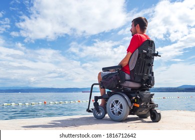 Man with muscular dystrophy on electric wheelchair outdoors looking at sea.