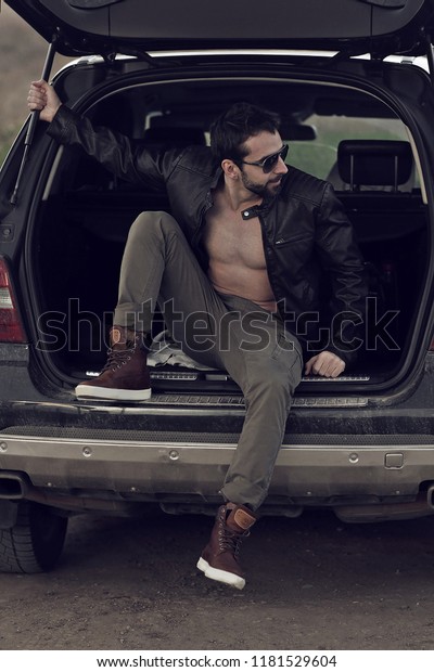 Man with muscular wet body and strong torso of\
bearded bodybuilder athlete in in a leather jacket with sunglasses\
on car trunk background