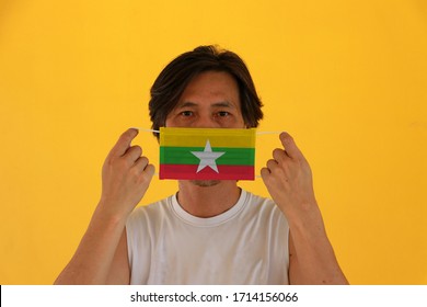 A Man With Myanmar Flag On Hygienic Mask In His Hand And Lifted Up The Front Face On Yellow Background. Tiny Particle Or Virus Corona Or Covid 19 Protection. Concept Of Combating Illness.