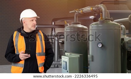 Man near heating equipment. Tanks for heating building. Basement with pipes and heating equipment. Guy controls boiler equipment. Man Engineer with phone. Worker in helmet in engineering room