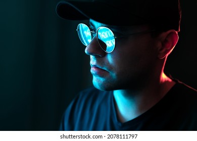 Man With Neon Light On Glasses And Face. Blue And Red Color From City Billboard In Dark Studio Portrait. Reflection In Sunglasses. Black Background In Dramatic Urban Night Portrait. Cyber Technology.