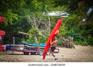 Man With No Shirt Struggling To Load Red Plastic Kayak On Float Trip Trailer On Pebbly River Beach With Lush Forest Background -selective Focus