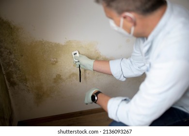 Man with nose mouth protection measures the moisture level on a wall with mold in an apartment