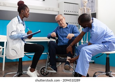 Man Nurse Checking Injured Knee Leg Of Sick Retired Senior Patient While Therapist Doctor Writing Medical Expertise On Clipboard During Clinical Examination In Hospital Office. Medicine Concept