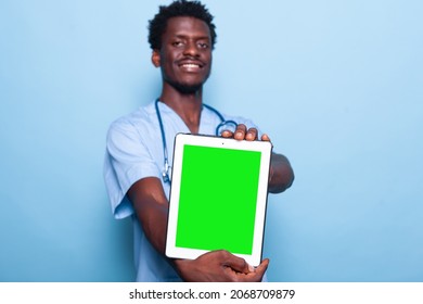 Man Nurse Holding Vertical Green Screen On Tablet In Studio. Medical Assistant Wearing Uniform And Stethoscope While Showing Gadget With Chroma Key And Isolated Mockup Template On Display.