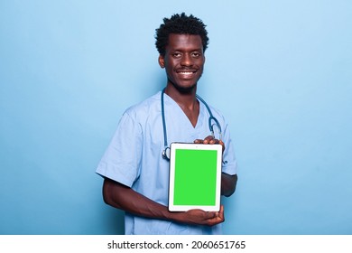 Man Nurse Showing Vertical Green Screen On Tablet. Medical Assistant Wearing Uniform And Stethoscope While Holding Device With Chroma Key And Isolated Mockup Template On Display.