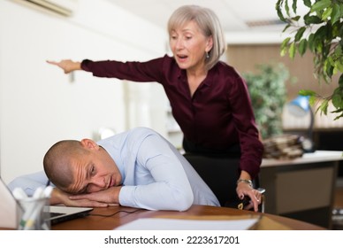 Man Office Worker Sleeping On Table During Work Day. His Chief Senior Woman Standing Next To Him And Shouting At Him.