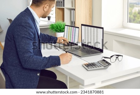 Man in office working with big data and databases using two laptops and excel tables. Office worker making analysis and report with spreadsheets on computer sitting at table near calculator.