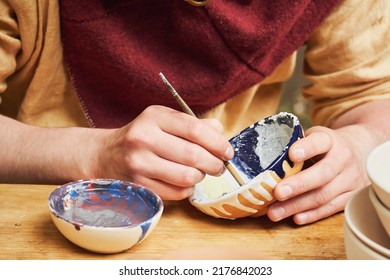 A Man In Old Medieval Byzantine Clothes Is Sitting Painting Clay Dishes With A Brush. Pottery In Nature In Retro Style.