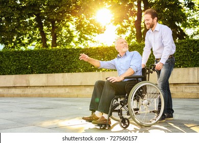 A man and an old man are walking in the park. The old man is sitting in a wheelchair. The man is carrying it