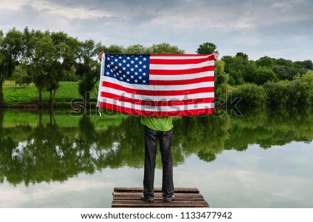 Man on the bank of the river on the shoulders with the flag of the USA. Having raised the flag up.