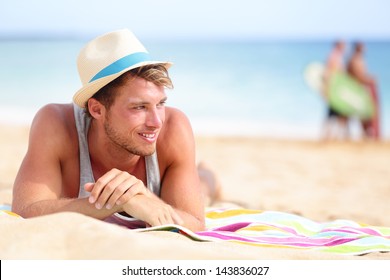 Man On Beach Lying In Sand Looking To Side Smiling Happy Wearing Hipster Summer Hat. Young Male Model Enjoying Summer Travel Holiday By The Ocean.