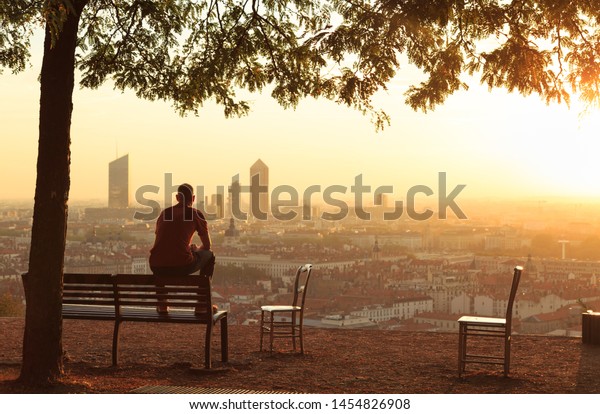 Man on a bench relaxing and enjoying the\
summer sunrise over a city. Lyon,\
France.