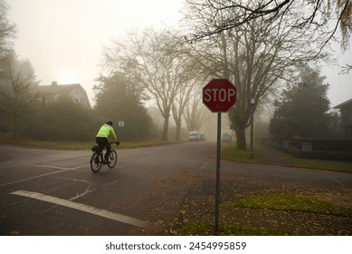 A man on a bicycle in a bright lime green jacket passing a deserted foggy intersection with a stop sign in an early autumn morning - Powered by Shutterstock