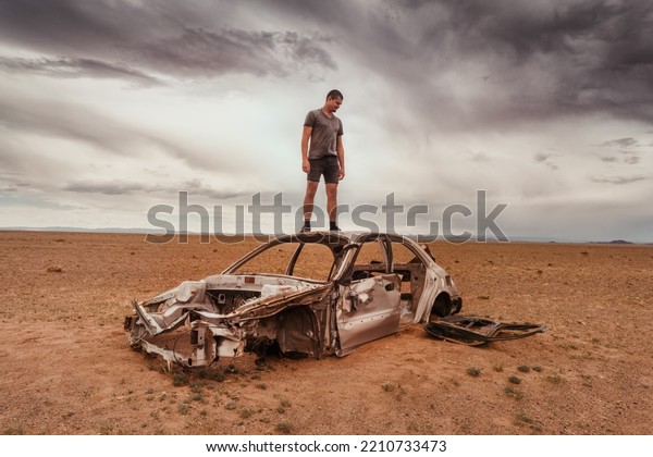 A man on a burnt-out car in the\
desert. A burnt-out abandoned car, under a grey gloomy\
sky