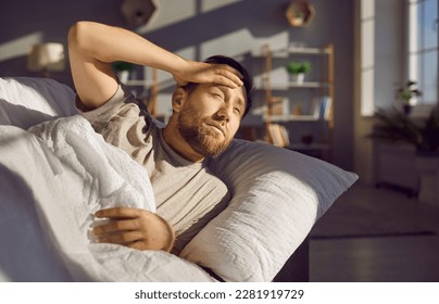 Man on the couch wakes up very early in the morning. Young man lying on the sofa in the living room at home and squinting in the morning sun with a sleepy face - Powered by Shutterstock