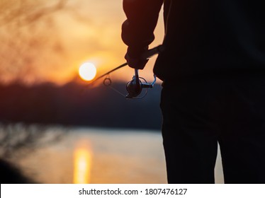 A Man On A Fishing Trip With A Spinning Rod Close-up Against The Background Of The River And Sunset