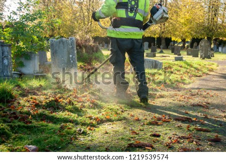 Man on his back working with vest and one gas string trimmer cutting the grass in a graveyard.