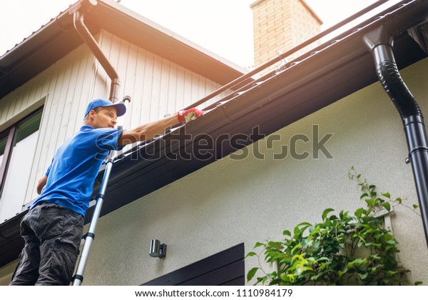 man\
on ladder cleaning house gutter from leaves and\
dirt