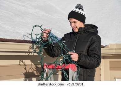 Man on a ladder putting up exterior Christmas lights on his house and trying to untangle the lights 