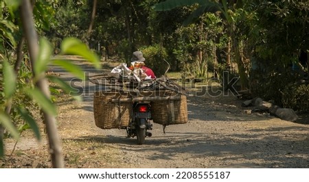 Man on a motorcycle on a rural road on the island of Bali, Indonesia. He is carrying a bicycle and two large baskets full of miscellaneous items. Indonesians carry large loads on their motor scooters