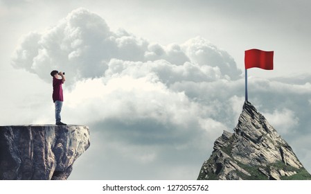 Man On A Mountain Peak Looking Through Binoculars To A Red Flag. The Concept Of Reaching Goals.