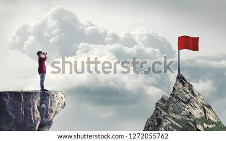 Man on a mountain peak looking through binoculars to a red flag. The concept of reaching goals.
