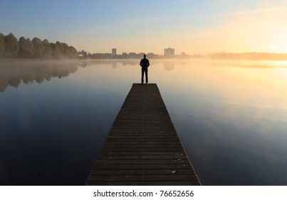 Man on a pier looking toward the city over a foggy lake during sunrise.