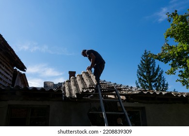 A Man On The Roof Of The House Repairs A Broken Roof Tile After A Big Storm On A Summer Day. Storm And Damage To Buildings Concept