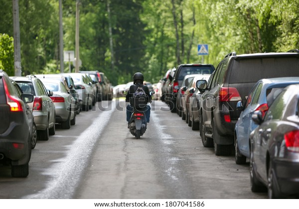 A\
man on a scooter drives off into the distance among parked cars\
against a background of green trees. Travel by\
Moto.