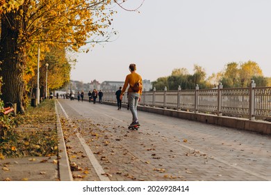 Man on skateboard on autumn promenade. A guy in yellow sweater with headphones rides a skateboard in alley on warm autumn day. Athletic man on city embankment of lake on a background of yellow leaves.