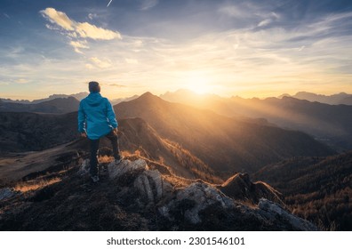 Man on stone on the hill and beautiful mountain valley in haze at colorful sunset in autumn. Dolomites, Italy. Guy, mountain ridges in fog, orange grass and trees, blue sky with sun in fall. Hiking	 - Shutterstock ID 2301546101
