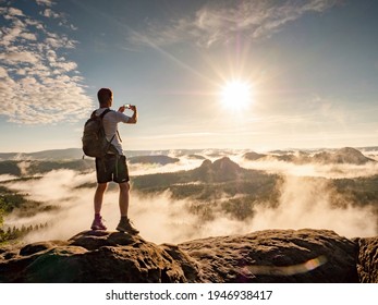 Man on top of a mountain watching the colorful sunset. Traveller with backpack photographing the beautiful nature scenics with smartphone while on vacation