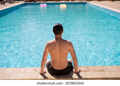 A Man On Vacation Sunbathes In Swimming Trunks Sitting By The Pool With Blue Water, Back View. Athlete With A Beautiful Body Relaxes On Vacation. Travel And Vacation Concept In Hotel. Place For Text